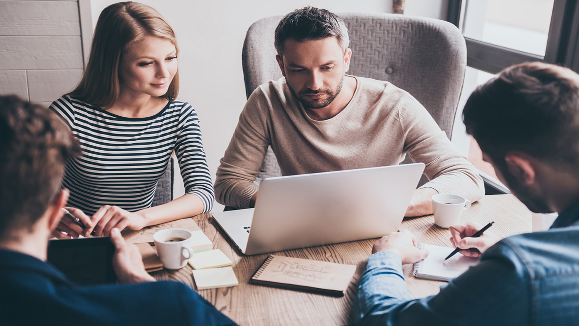 Redaktionsmöte med fyra personer vid ett bord med datorer, papper, en av dem antecknar